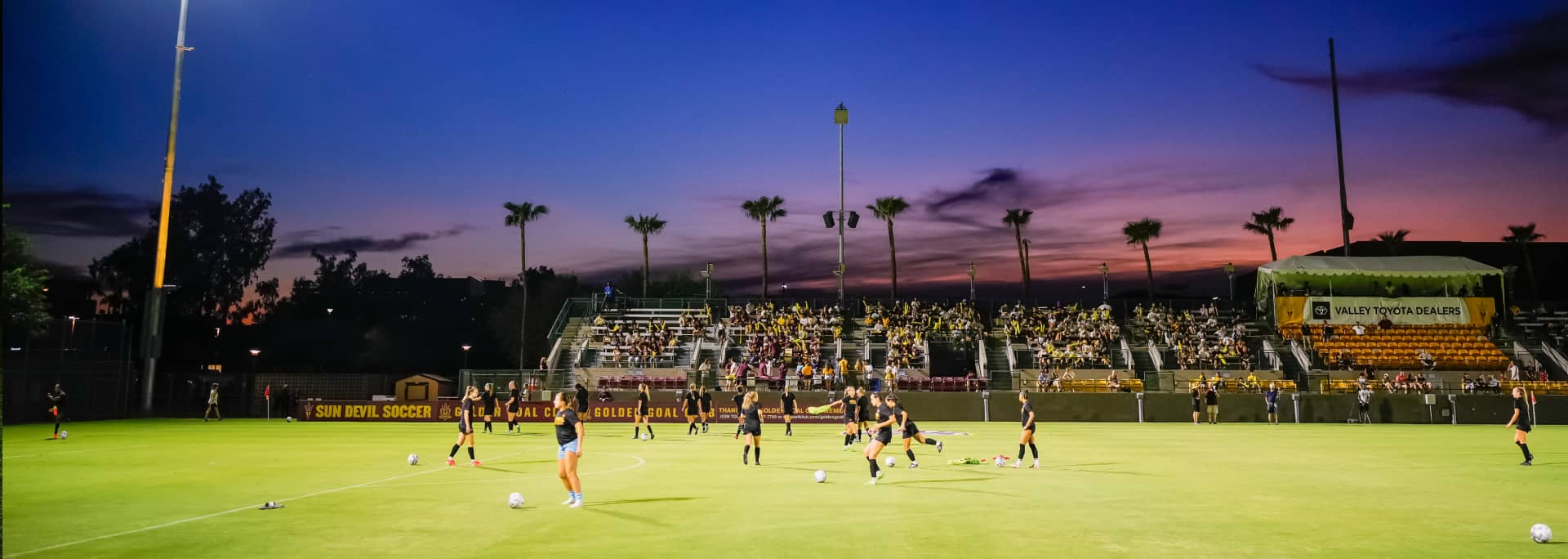 A zoomed out image of the ASU soccer field. ASU soccer players are practicing while people watch from the stands and the sun is setting in the distance.