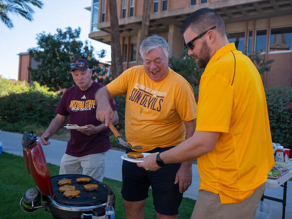 Three men dressed in ASU t-shirts grilling hamburgers outside.